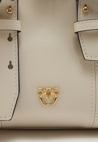 Krémová kožená kabelka s zlatými kovovými detailmi v tvare vtáka, kontrastným tmavým prešívaním a štruktúrovaným dizajnom s prackovými akcentmi.