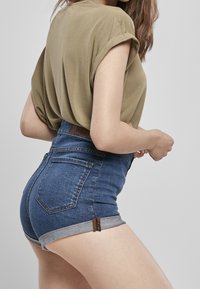 Højtaljede denim shorts med opfoldede kanter, mørkeblå vask, to baglommer og et lædermærke ved taljen, parret med en løs olivengrøn top.