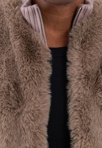 Taupe faux fur jacka med hög krage, med en ribbad ljusrosa accent och silverfärgad dragkedja. Texturerad, mjuk look. Svart ribbad topp under.
