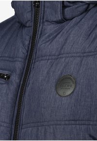 Polstret jakke i mørkeblått stoff med teksturert overflate, med en svart glidelås og et rundt svart logo-patch på brystet.