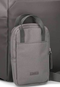 Bolsa de tela gris con doble asa, forma rectangular, cierre con cremallera, bolsillo frontal y logo grabado. Superficie texturizada y costuras limpias.