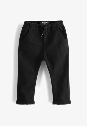 Tout-petit portant un t-shirt rayé gris et blanc, un pantalon noir et des chaussures blanches, marchant sur fond blanc uni.