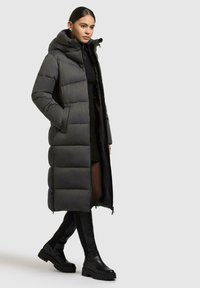 Lungo cappotto imbottito grigio con cappuccio, caratterizzato da un design trapuntato e tasche con zip, indossato su un outfit nero e abbinato a stivali neri al ginocchio.