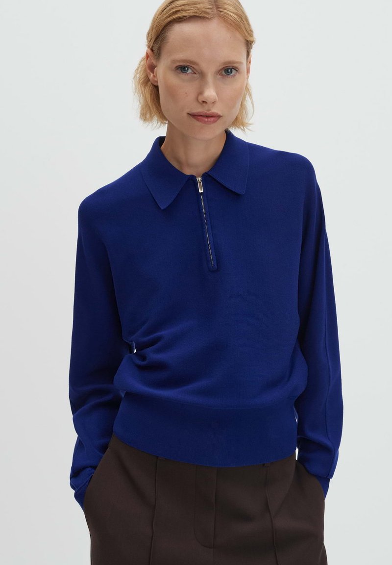 Pull-over en maille bleue avec col à zip, manches longues et ourlet ajusté. Texture douce ; porté avec un pantalon marron taille haute.
