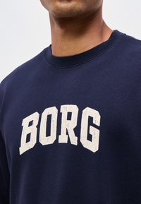 Homme portant un sweat-shirt bleu marine avec des lettres majuscules blanches formant "BORG" sur la poitrine.