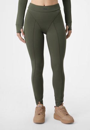 Frau trägt olivgrüne High-Waist-Leggings mit sichtbaren Nahtdetails und beigen, klobigen Sneakers mit Logo-Branding.