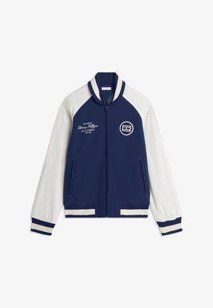 Veste zippée bleu marine et blanche avec manches blanches, poignets et col côtelés rayés, poches avant, texte et logo brodés sur la poitrine.