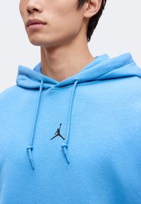 Gros plan d'une personne portant un sweat à capuche bleu avec un logo noir de joueur de basketball sur la poitrine et des cordons pendant.