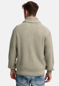 Suéter de punto gris claro con un patrón texturizado de waffle, cuello chal y puños y dobladillo en canalé. Usado sobre jeans azul claro.