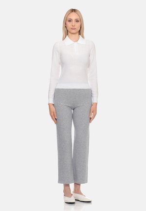 Donna bionda con indosso una maglia bianca a manica lunga con colletto, pantaloni grigi a gamba larga e scarpe basse bianche, in piedi davanti a uno sfondo semplice.