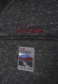 Black knitted fabric with a speckled texture. Features red embroidered logo and a colorful patch with mountain design labeled “POLAR EXPERIENCE 84.”