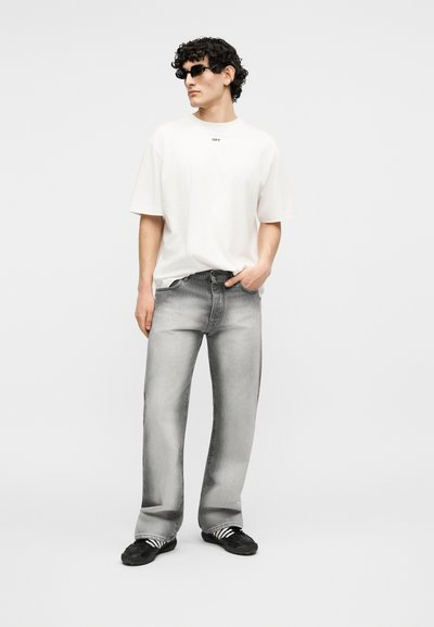 Άνδρας με μαύρα σγουρά μαλλιά που φοράει μαύρα γυαλιά ηλίου, λευκό φαρδύ μπλουζάκι, ξεθωριασμένο γκρι τζιν και μαύρα αθλητικά παπούτσια με λευκές ρίγες, όρθιος.