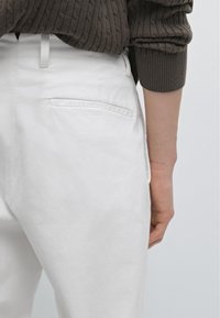 Witte katoenen broek met een gladde textuur, met een achterzak en ceintuurlussen. De zichtbare hand rust op de tailleband.