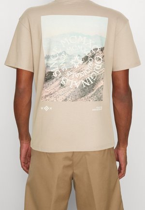 Camiseta de algodón beige con un gran gráfico de paisaje montañoso y diseño de texto circular en la parte posterior. Mangas cortas, corte relajado.