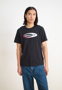 Fekete pamut póló, fehér és piros ovális logóval, amelyen a "Reebok" márkaneve szerepel. Rövid ujjak és klasszikus kerek nyakú kialakítás.