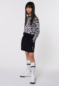 Sweatshirt graphique noir et blanc avec logo DKNY, associé à un short noir. Chaussettes blanches avec rayures noires et baskets montantes blanches.
