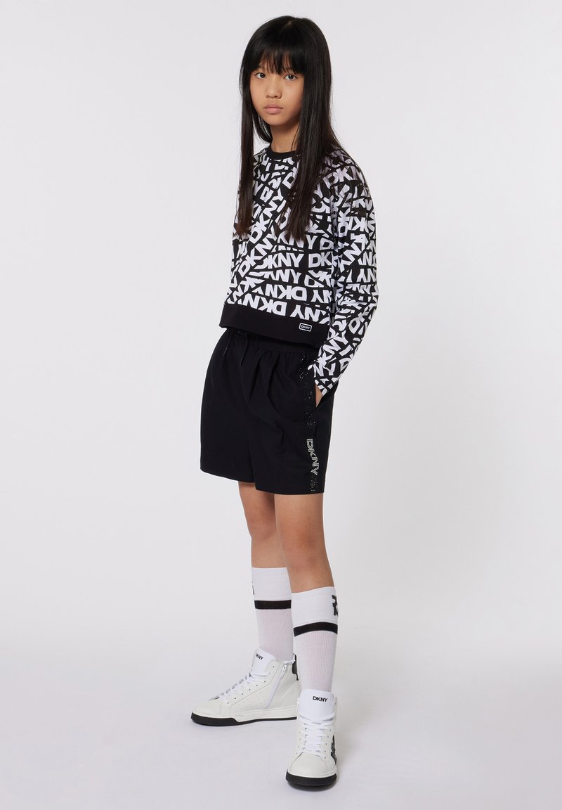 Sweatshirt graphique noir et blanc avec logo DKNY, associé à un short noir. Chaussettes blanches avec rayures noires et baskets montantes blanches.