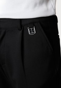Black trousers made of textured fabric, featuring a silver logo buckle and front pleats. The waistband lies beneath a white ribbed top.