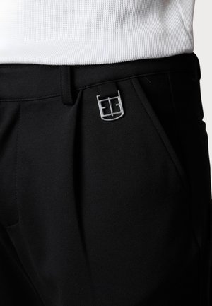 Black trousers made of textured fabric, featuring a silver logo buckle and front pleats. The waistband lies beneath a white ribbed top.