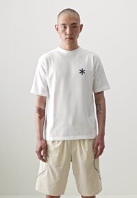 T-shirt blanc à manches courtes avec un symbole d'étoile noir sur la poitrine gauche. Le modèle l'associe avec un short beige, mettant en évidence une coupe décontractée.