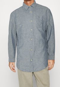 Button-up shirt in light blue fabric with white vertical stripes. Features a classic collar, long sleeves, and a chest pocket.