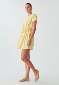 Geltona kasdienė suknelė su trumpomis rankovėmis, suplatinta juosmeniu ir plazdančia sijonu. Nešiojama su plokščiais sandalais, turinčiais plonus dirželius.