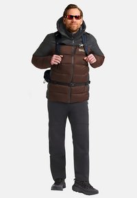 Gevoerde donsjas in donkerbruin en grijs met ritssluiting, verstelbare capuchon en voorzakken; gecombineerd met een donkere broek en wandelschoenen.