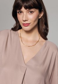 Złoty naszyjnik łańcuszkowy z splecionymi ogniwami, noszony na lekko różowej, miękkiej bluzce z dekoltem w kształcie litery V. Gładka faktura, nowoczesny wygląd.