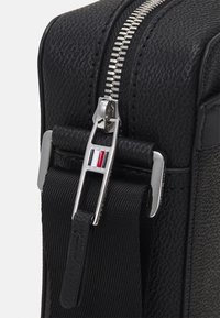 Borsa a tracolla nera con texture, dotata di cerniera e accessori in argento. Tracolla regolabile con un accento in pelle e un dettaglio rosso, bianco e blu sul tirante della cerniera.