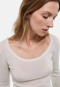 Haut à manches longues léger et côtelé en blanc doux avec un large décolleté et une silhouette ajustée. La peau est visible à travers le tissu transparent.