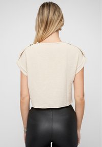 Top beige cropped con maniche arrotolate e dettagli con bottoni. La texture stropicciata conferisce un aspetto informale, abbinato a leggings neri aderenti.