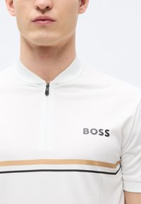 Λευκό κοντομάνικο πόλο με γιακά μισής φερμουάρ. Διαθέτει οριζόντιες ρίγες σε μαύρο και χρυσό και λογότυπο "BOSS" σε μαύρο στο στήθος. Ομαλή υφή.