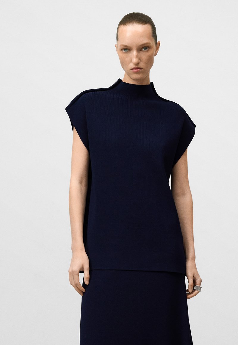 Top lavorato a maglia navy con colletto alto, maniche a sbuffo e vestibilità rilassata. Tessuto liscio con dettagli di cucitura sottili. Abbinato a una gonna coordinata.