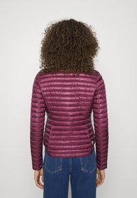 Lätt, plommonfärgad pufferjacka med horisontell quiltning, slät textur och åtsittande design. Har en logotyp på nedersta vänstra sidan.