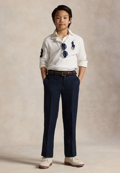 Fehér hosszú ujjú póló sötétkék logóval, sötétkék nadrág, fonott barna öv, és világos színű cipők semleges háttéren.