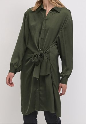 Olijfgroene blousejurk met lange mouwen en knopen, met een strik aan de voorkant ter hoogte van de taille, gedragen over een zwarte broek door een staand persoon met blond haar.