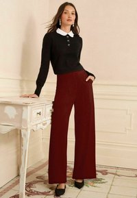 Melns džemperis ar baltu apkakli un dekoratīvām pogām, kombinēts ar augstu jostasvietu platām bordo biksēm. Vienkāršs dizains un eleganta piegrieztnes.