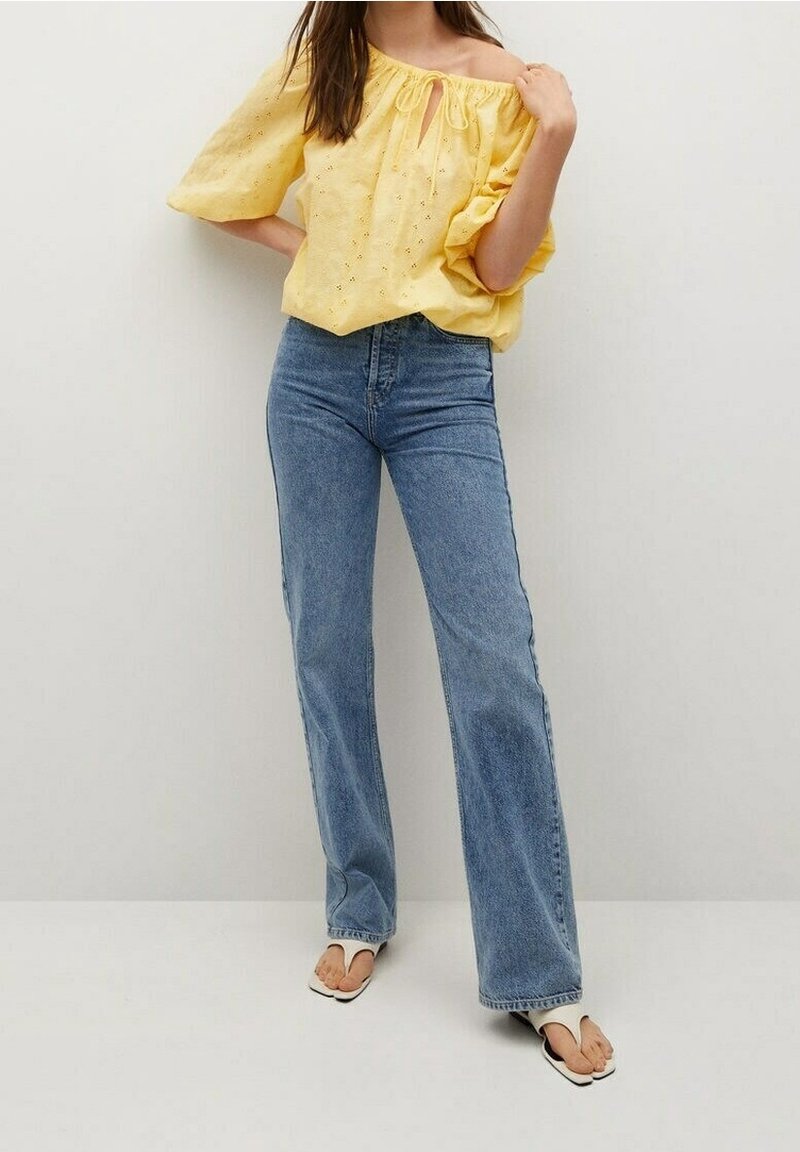 Femme portant un chemisier jaune à épaules dénudées, un jean bleu taille haute et des sandales blanches, debout devant un fond clair uni.