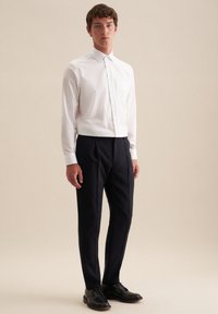 Camicia bianca con bottoni e maniche lunghe, taschino sul petto; abbinata a pantaloni blu scuro e scarpe nere; design semplice, texture liscia.