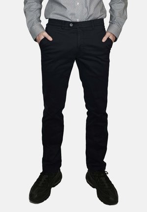 Pantaloni neri slim fit in cotone con una texture liscia. Tasche frontali, chiusura con bottoni, abbinati a scarpe sportive nere.