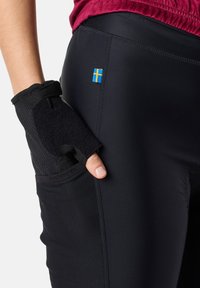 Schwarze Sporthosen verfügen über einen strukturierten Abschnitt an den Seiten und ein kleines schwedisches Flaggenakzent. Eine Hand mit einem schwarzen Fingerloshandschuh liegt in einer Tasche.