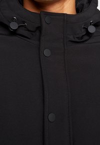 Gros plan sur une veste noire avec des boutons-pression et des cordons de capuche réglables sur la partie supérieure de la poitrine et le cou d'une personne.