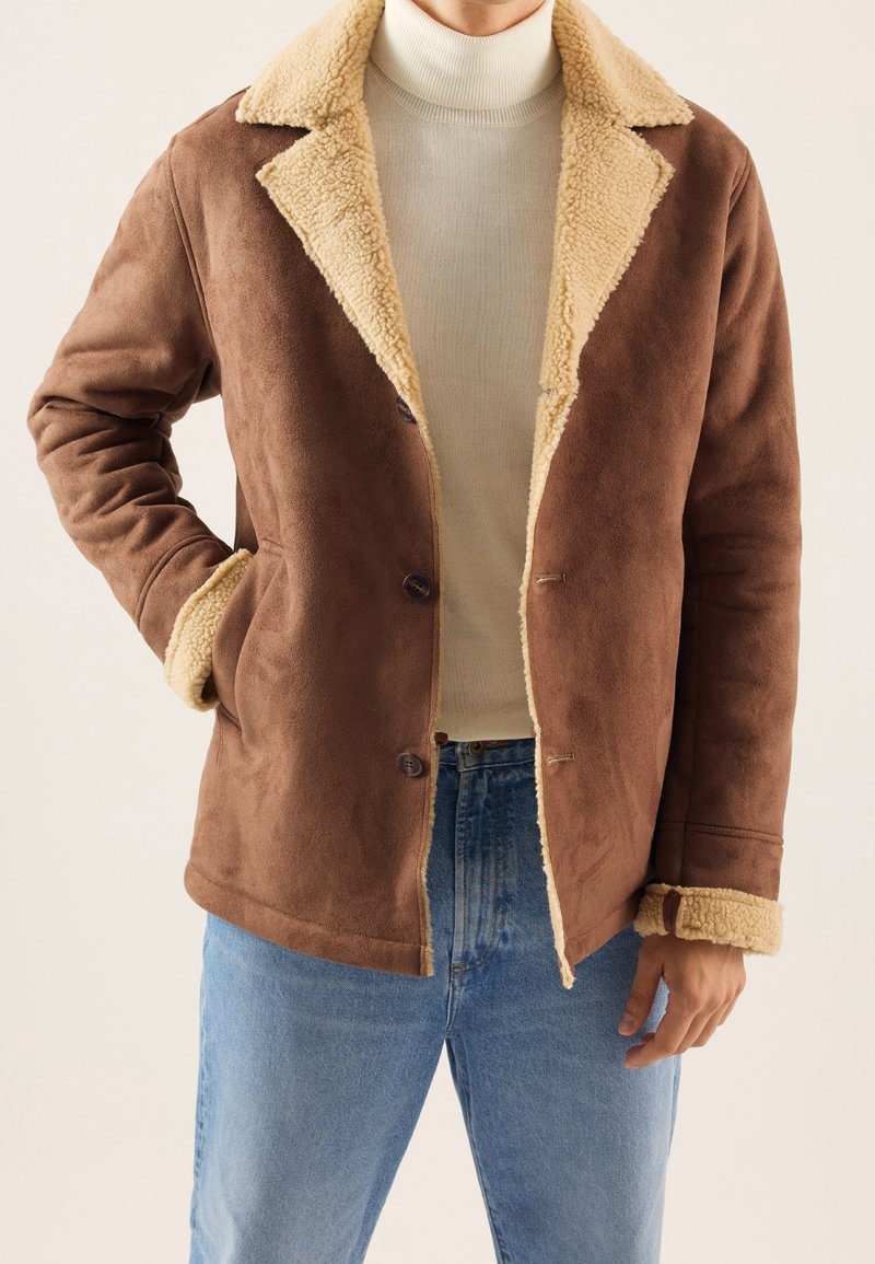 Homme portant une veste en peau retournée marron avec doublure en polaire crème, un pull à col roulé beige et un jean bleu, sur un fond neutre.