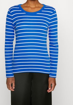 Personne portant une chemise bleue à manches longues avec des rayures blanches horizontales et un pantalon noir, debout devant un fond clair uni.