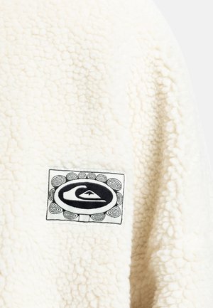 Tissu en polaire crème avec une surface texturée ; présente un logo noir brodé sur une patch rectangulaire.