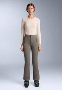 Beiger Langarmtop mit einem strukturierten Diamantmuster, kombiniert mit eng anliegenden grauen Hosen mit hoher Taille und vorderen Reißverschlusstaschen. Schwarze Skistiefel.