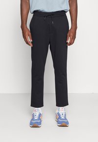 Zwarte trainingsbroek met een elastische tailleband en drawstring. De stof lijkt zacht; de broek loopt iets taps toe bij de enkels. Gedragen met blauwe sportschoenen.