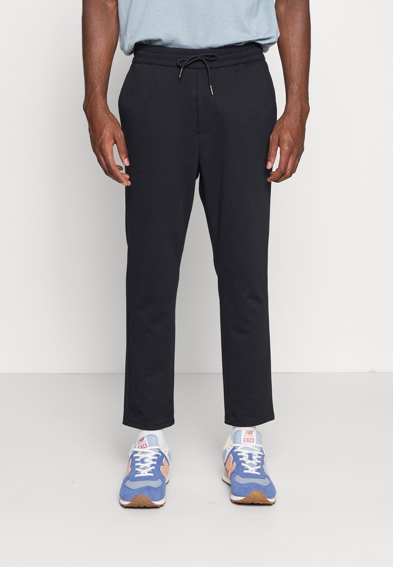 Zwarte trainingsbroek met een elastische tailleband en drawstring. De stof lijkt zacht; de broek loopt iets taps toe bij de enkels. Gedragen met blauwe sportschoenen.