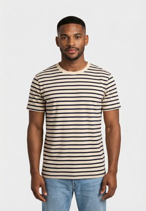 Uomo con i capelli corti e la barba che indossa una maglietta a righe orizzontali beige e nera e jeans azzurri, in piedi con le braccia rilassate.