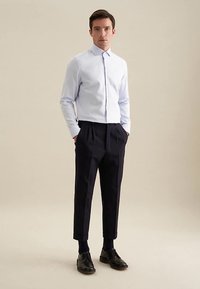 Lichtblauw overhemd met lange mouwen, slim fit, met knoopsluiting op de kraag. Donkere geplooide broek, cropped lengte, gedragen met zwarte schoenen en navy sokken.